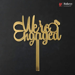 Gold metal 'We are Engaged' cake topper with a ring icon, shown on a black background with a Bakeey brand watermark in the top right corner.