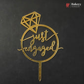 A gold metal 'just engaged' cake topper with a diamond graphic and stake, displayed on a dark textured background. A Bakeyy logo is visible in the top right corner.