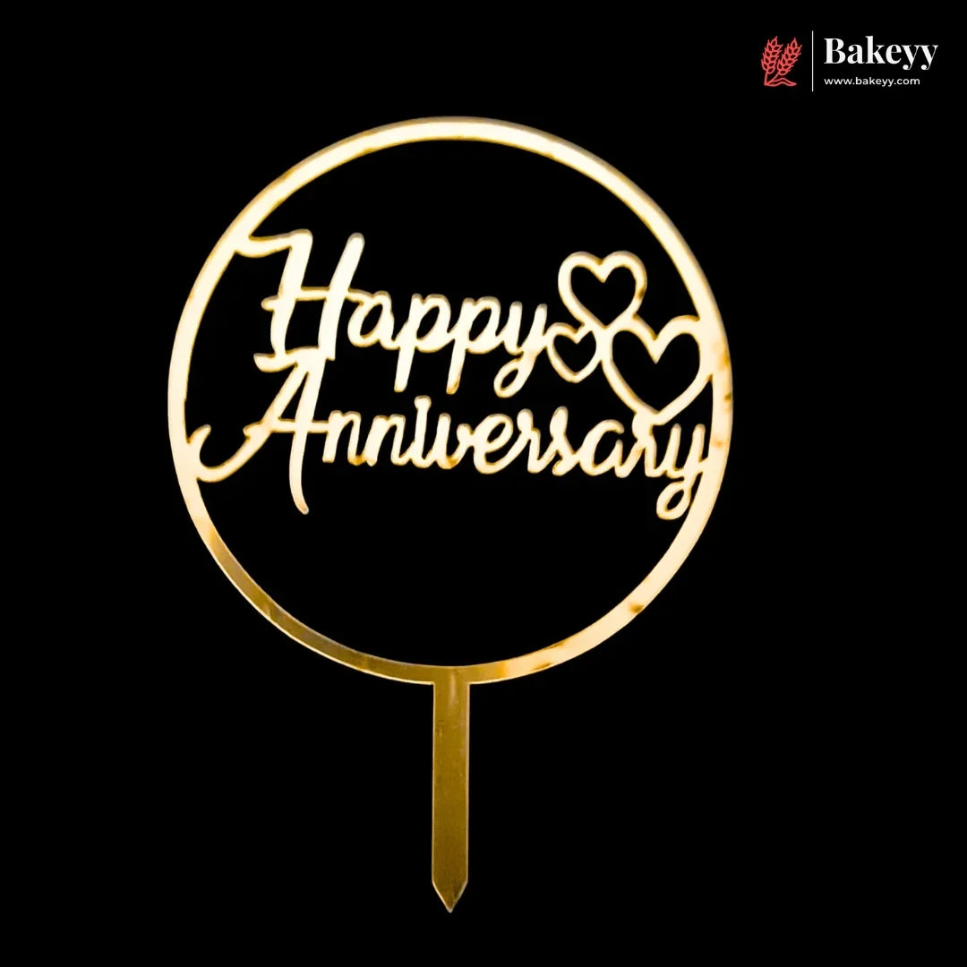A gold-colored metal cake topper with a circular frame and stake, featuring the words 'Happy Anniversary' in cursive script with two heart shapes, shown against a solid black background. A 'Bakeyy' brand watermark is visible in the top right corner.