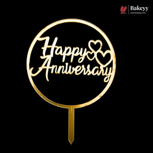 A gold-colored metal cake topper with a circular frame and stake, featuring the words 'Happy Anniversary' in cursive script with two heart shapes, shown against a solid black background. A 'Bakeyy' brand watermark is visible in the top right corner.