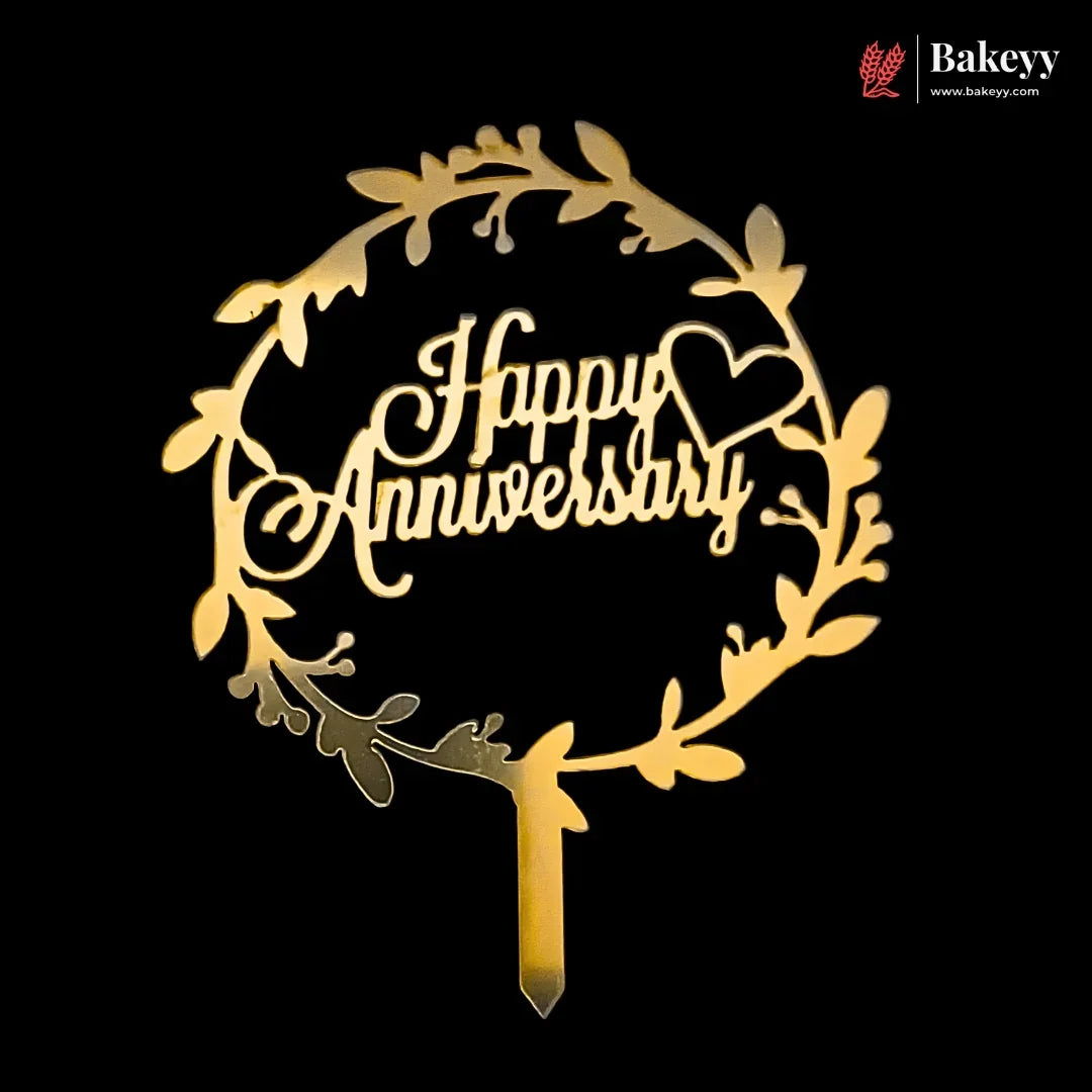 A gold-colored wooden cake topper with a circular leaf wreath design and the words 'Happy Anniversary' in cursive script, shown on a solid black background with a Bakeey brand watermark in the top right corner.