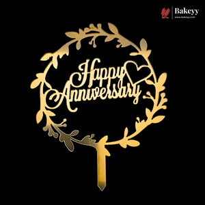 A gold-colored wooden cake topper with a circular leaf wreath design and the words 'Happy Anniversary' in cursive script, shown on a solid black background with a Bakeey brand watermark in the top right corner.