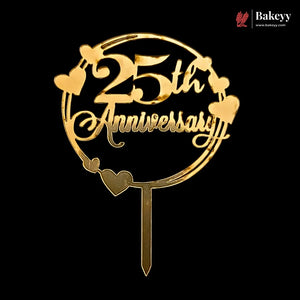 A gold-colored metal cake topper featuring the text '25th Anniversary' inside a circular frame with heart accents, shown on a solid black background. A Bakeyy brand logo is visible in the top right corner.