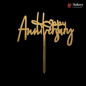 Gold acrylic 'Happy Anniversary' cake topper with a metal wire stand, shown on a solid black background. A Bakeyy brand logo is visible in the top right corner.