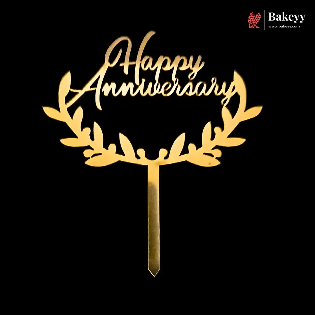 Gold laser-cut acrylic cake topper with 'Happy Anniversary' script and leafy wreath design on a black background, with a Bakeyy logo in the top right corner.