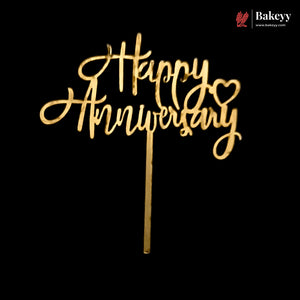 Gold-colored wooden 'Happy Anniversary' cake topper with a heart detail and a single stick, shown against a solid black background. A 'Bakeyy' brand logo is visible in the top right corner.