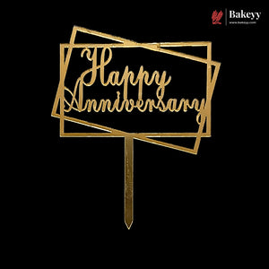 Gold metal 'Happy Anniversary' cake topper with a geometric frame design and stake, shown on a solid black background. A Bakeyy logo is visible in the top right corner.
