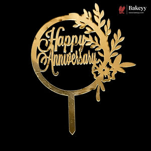 A gold-colored metal 'Happy Anniversary' cake topper with a leafy wreath design and a stake, shown against a solid black background. A Bakeyy brand logo is visible in the top right corner.