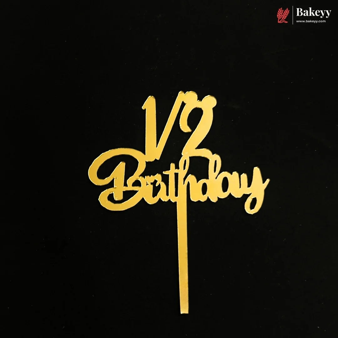 Gold metal cake topper with '1/2 Birthday' script on a black background, featuring a Bakeyy brand logo in the top right corner.
