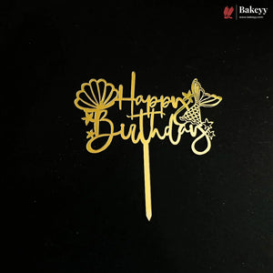 A gold metal cake topper with 'Happy Birthday' script, decorated with a seashell and butterfly design, shown on a black background with a Bakeyy brand watermark in the top right corner.