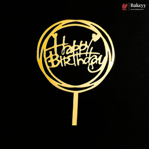 A gold metal 'Happy Birthday' cake topper with a circular frame and stick base, shown against a solid black background. A Bakeey brand logo is visible in the top right corner.