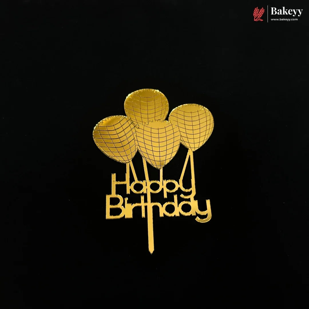 A gold-colored metal cake topper featuring three balloon shapes above the words 'Happy Birthday' in cursive, displayed on a solid black background. A 'Bakeyy' brand logo is visible in the top right corner.