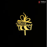A gold metal cake topper shaped like a gift box with a bow and the words 'Happy Birthday' on a black background. A small Bakeey brand logo is visible in the top right corner.