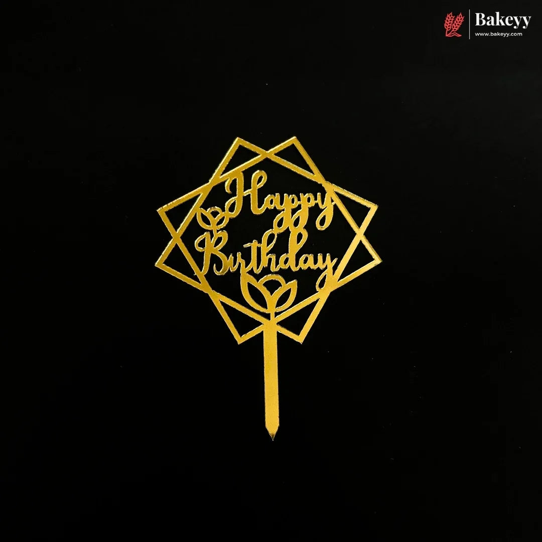 A gold metal cake topper with a geometric diamond frame and the words 'Happy Birthday' in cursive, displayed on a solid black background. A Bakeyy logo is visible in the top right corner.