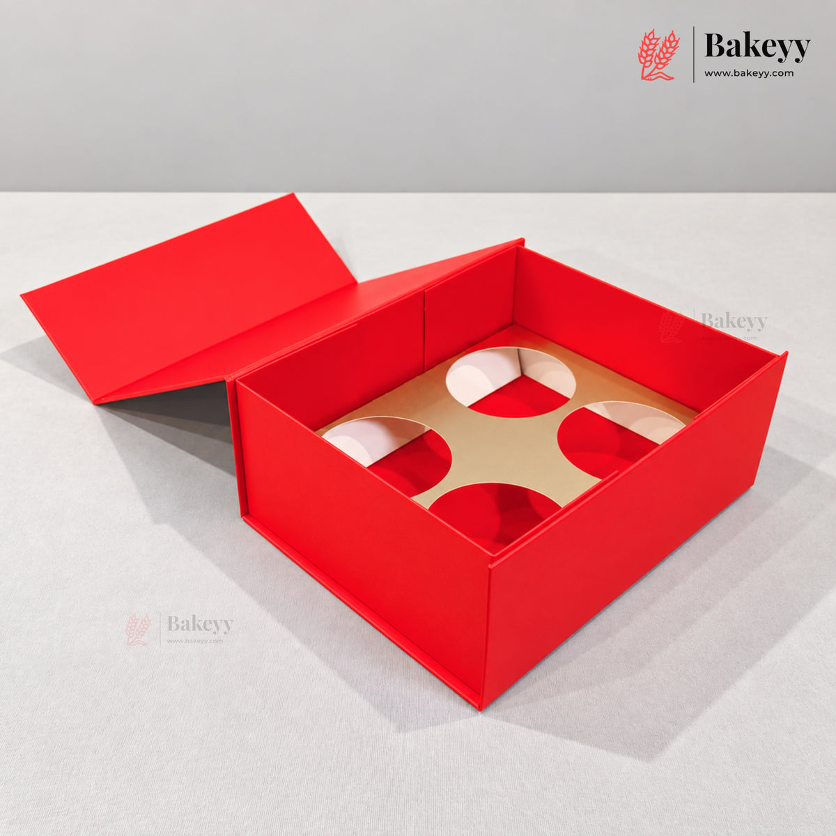 A red bakery gift box with a hinged lid open, showing a tan cardboard insert with four circular cutouts, photographed on a light gray surface against a gray background. The Bakeyy logo appears in the top right corner.