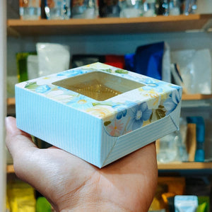 A hand holding a square floral gift box with a clear window on top, set against a blurred background of shelves and items.