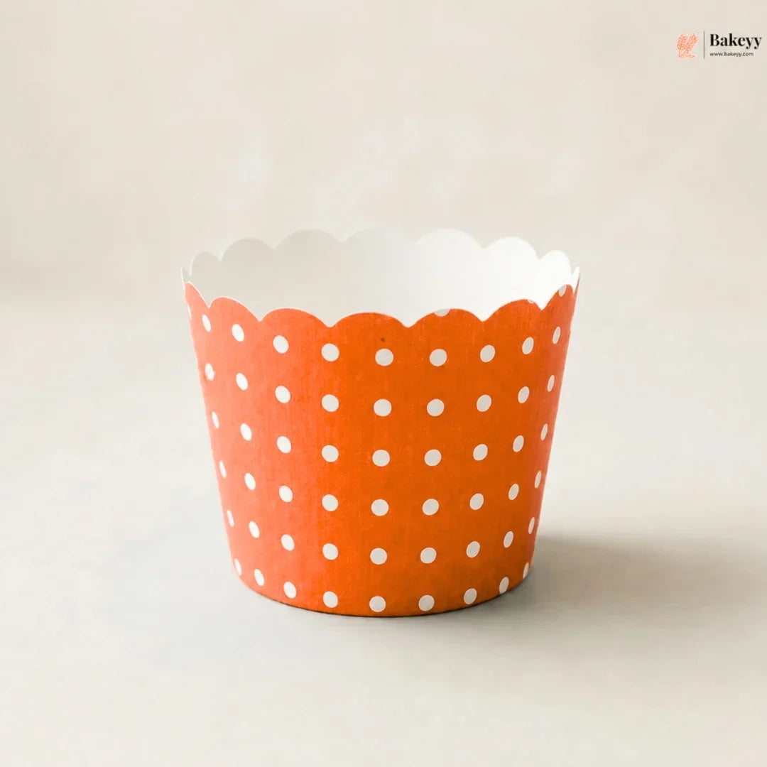 An orange paper cupcake liner with a white polka dot pattern and scalloped edge, photographed on a neutral light beige background.