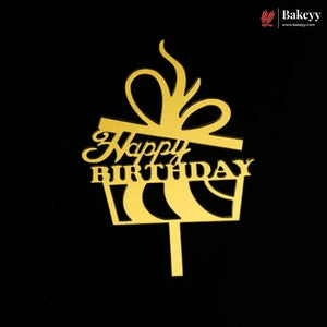 A gold metal cake topper shaped like a gift box with a bow and the words 'Happy Birthday' in script, shown on a black background. The Bakeey brand logo is visible in the top right corner.
