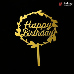 A gold metal cake topper with a circular leaf wreath and the words 'Happy Birthday' in script, shown on a black background. A Bakeey logo is visible in the top right corner.