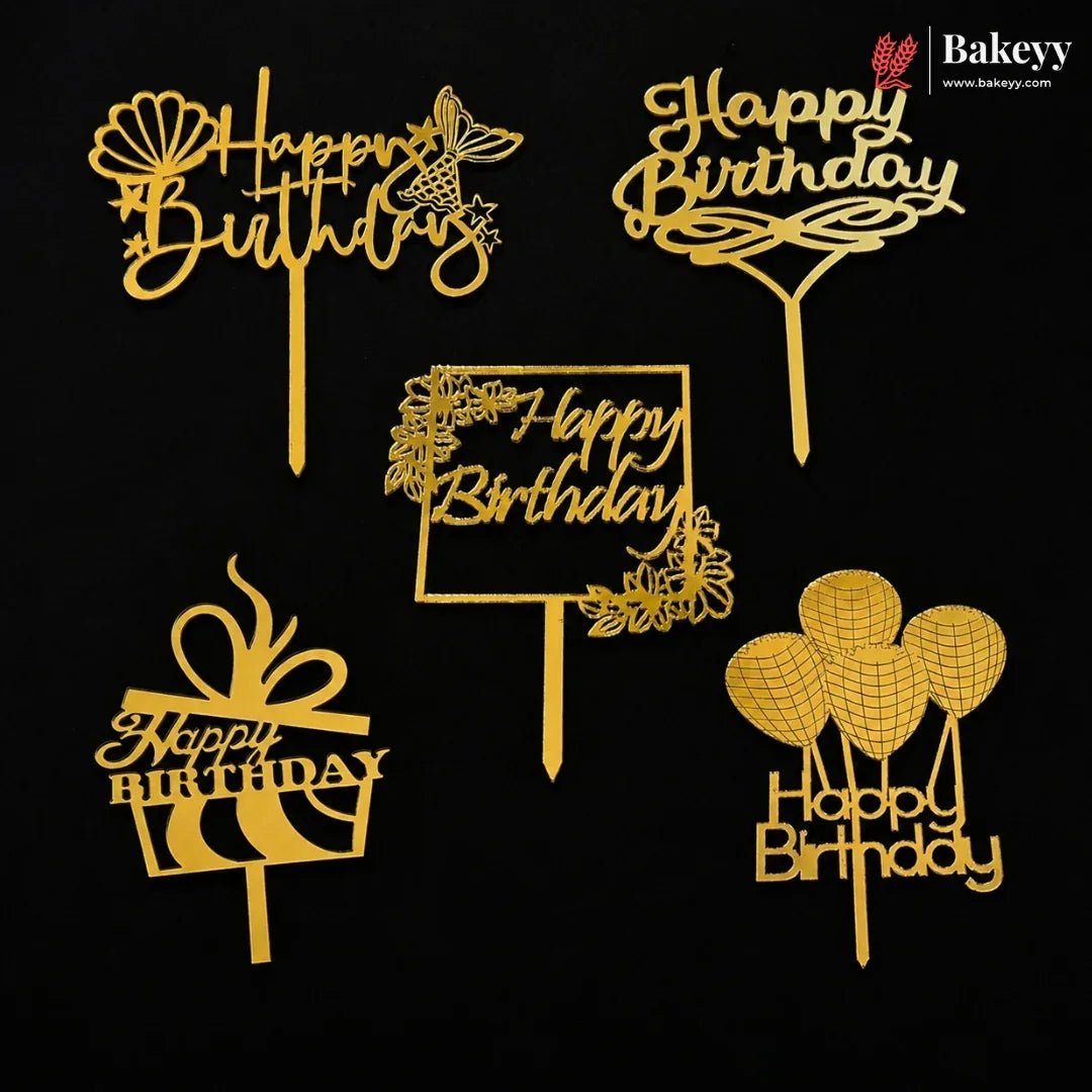 Six gold-colored plastic 'Happy Birthday' cake toppers in various designs, including a gift box, balloons, flowers, and a sign, arranged on a solid black background. A 'Bakeey' brand logo is visible in the top right corner.