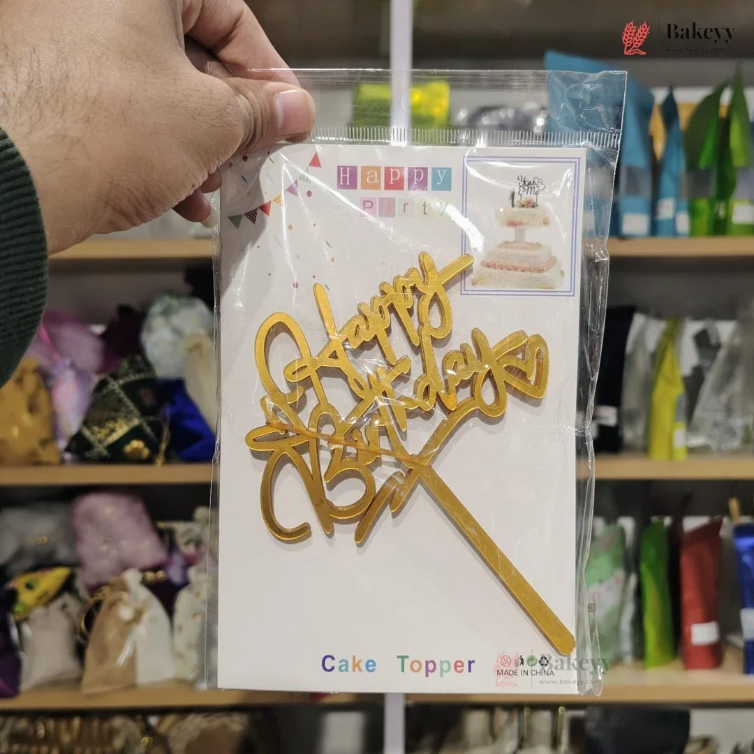 Gold 'Happy Birthday' cake topper in packaging held by a person with a blurred store background.