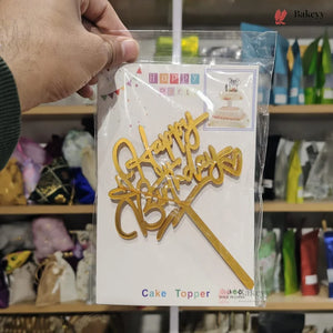 Gold 'Happy Birthday' cake topper in packaging held by a person with a blurred store background.