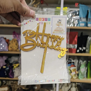 Gold 'Happy Birthday' cake topper in packaging held by a hand with a store shelf in the background.