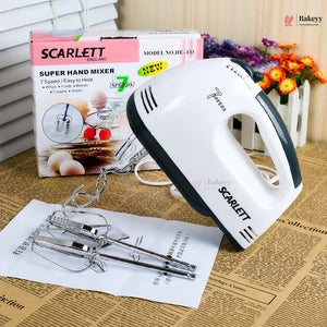 Scarlett hand mixer with attachments on a newspaper, next to its packaging and flowers in the background.
