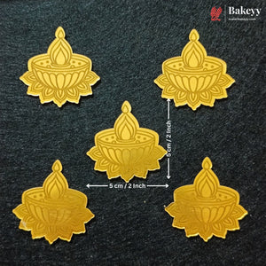 Five gold-colored metal oil lamp molds arranged on a dark textured background, with dimension arrows and text indicating a size of 5 cm by 5 cm (2 inches) around the central piece. A Bakeyy logo is visible in the top right corner.