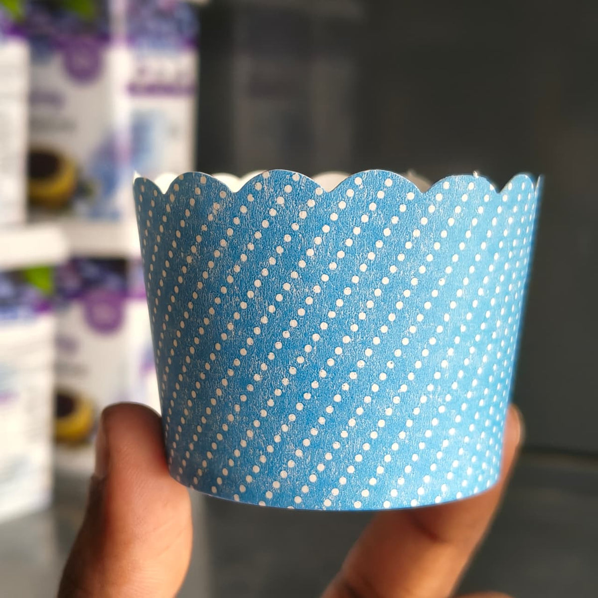 A hand holding a blue paper cupcake liner with a white polka dot pattern, with blurred packaged goods in the background.