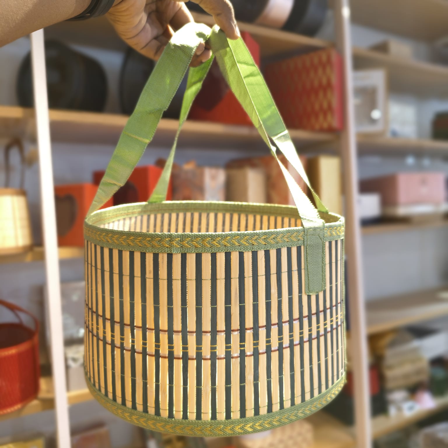 A hand holds a round woven basket with green fabric handles and a striped bamboo body, set against a blurred shop background.