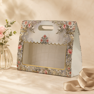 A decorative paper gift box with a floral print, scalloped window trim, and a handle, photographed on a warm beige surface with a vase of flowers and a ribbon nearby.