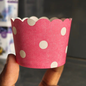A pink paper cupcake liner with white polka dots and a scalloped edge, held by a hand against a blurred dark background.