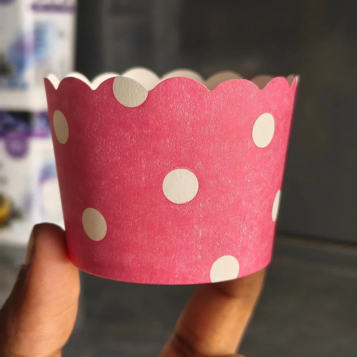 A pink paper cupcake liner with white polka dots and a scalloped edge, held by a hand against a blurred dark background.