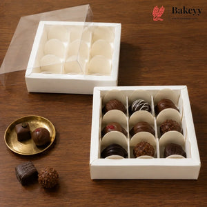 A white square chocolate box with a clear lid, shown open with multiple compartments holding assorted chocolates. A second empty compartment section of the same box is shown behind it, and two chocolates rest on a small gold plate beside the main box on a wooden surface.