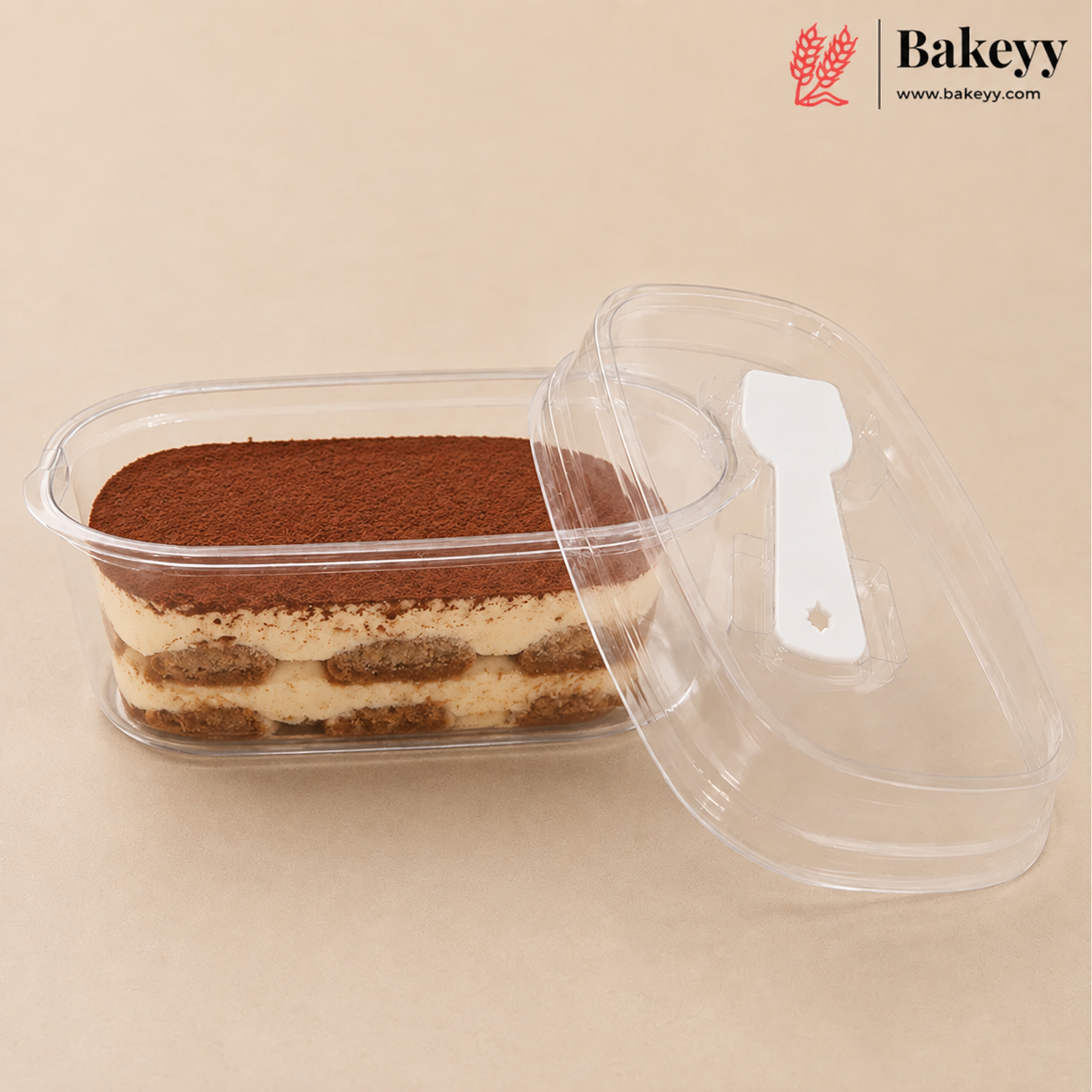 A clear plastic rectangular food container holding a layered tiramisu dessert, with a matching clear lid leaning beside it on a beige background. The Bakeyy logo appears in the top right corner.