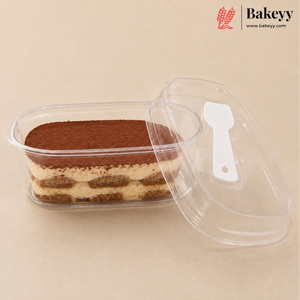 A clear plastic rectangular food container holding a layered tiramisu dessert, with a matching clear lid leaning beside it on a beige background. The Bakeyy logo appears in the top right corner.