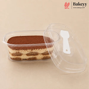 A clear plastic rectangular food container holding a layered tiramisu dessert, with a matching clear lid leaning beside it on a beige background. The Bakeyy logo appears in the top right corner.