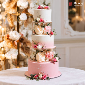 A five-tier pastel pink and white wedding cake decorated with pink and cream roses in clear square boxes, displayed on a white tablecloth with a blurred floral background. The Bakeey brand logo is visible in the top right corner.