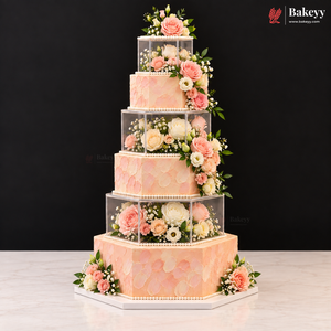 A five-tier peach and pink floral cake with clear acrylic cube risers, displayed on a white marble surface against a black background. The Bakeyy logo is visible in the top right corner.