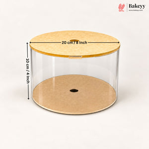 A clear cylindrical cake or pastry display cylinder with a light wood lid and a kraft paper base, shown on a white background with dimension arrows indicating 20 cm / 8 inch in diameter and 10 cm / 4 inch in height. Bakeyy logo is visible in the top right.