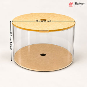 Clear cylindrical cake display box with a light wood lid and base, shown on a white background with dimension arrows and text indicating 25 cm / 10 inch diameter and 11.5 cm / 4.
