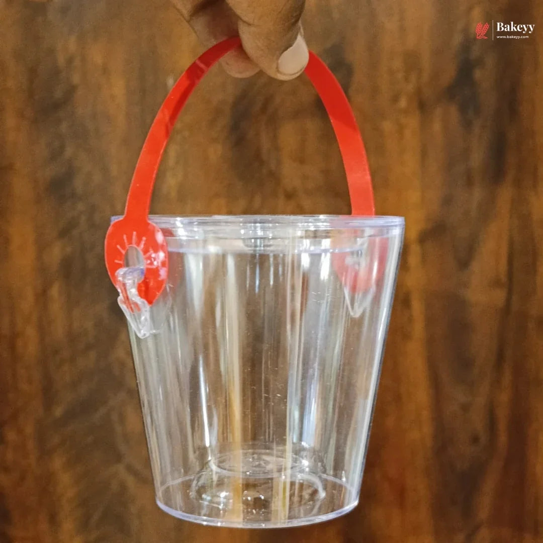 Clear plastic bucket with a red handle held by a hand against a wooden background