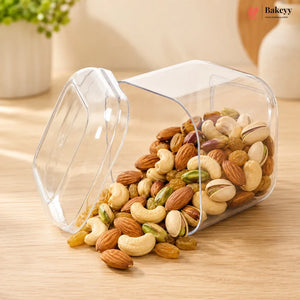A clear plastic cylindrical storage container with a hinged lid, tipped over on a light wood table with mixed nuts and raisins spilling out. A Bakeyy logo is visible in the top right corner.