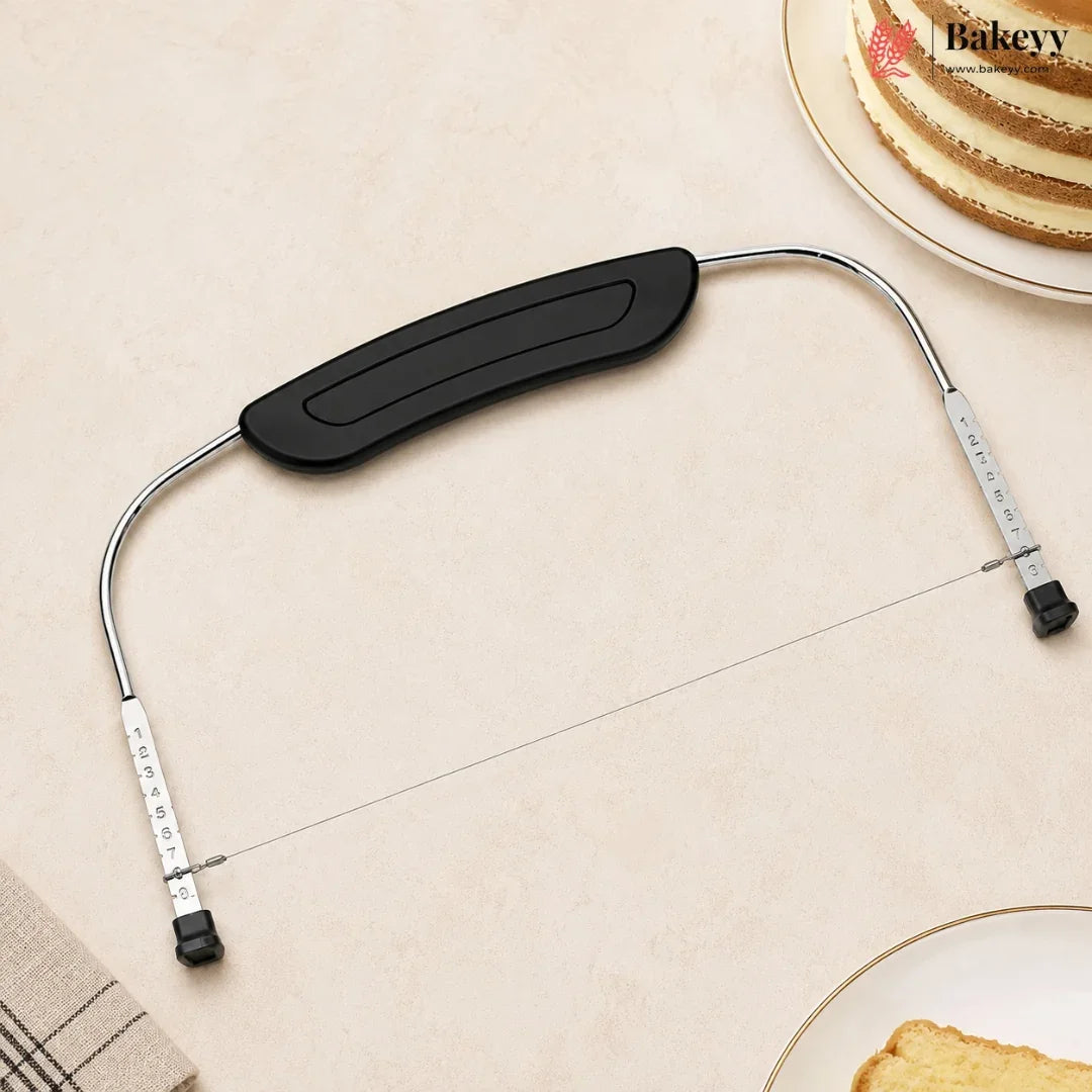 A metal bakery tool with a black curved handle and silver wire, laid on a beige surface with partial plates of cake visible in the corners. The brand name Bakeyy appears in the top right.