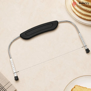 A metal bakery tool with a black curved handle and silver wire, laid on a beige surface with partial plates of cake visible in the corners. The brand name Bakeyy appears in the top right.