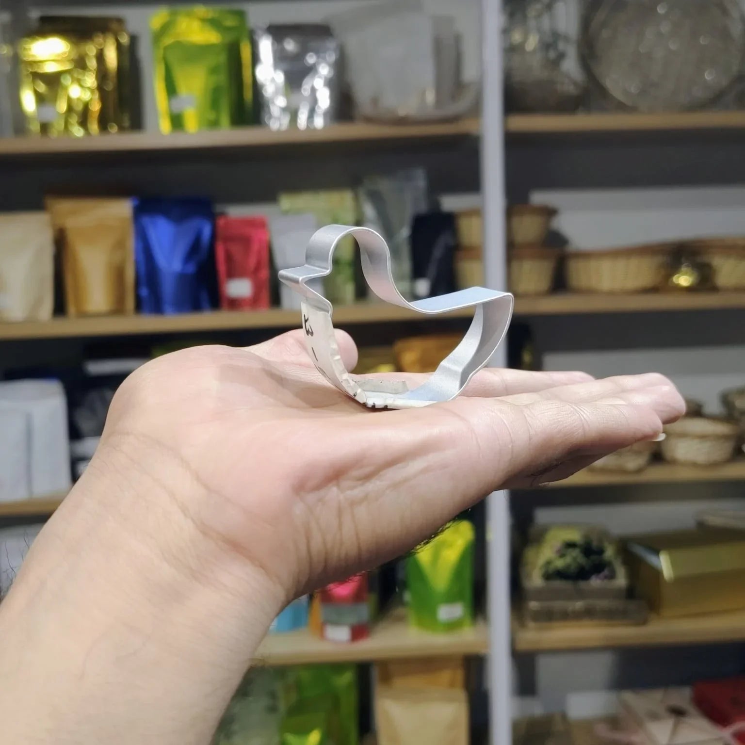 Hand holding a small metal cookie cutter shaped like a fish in front of a shelf with various items.