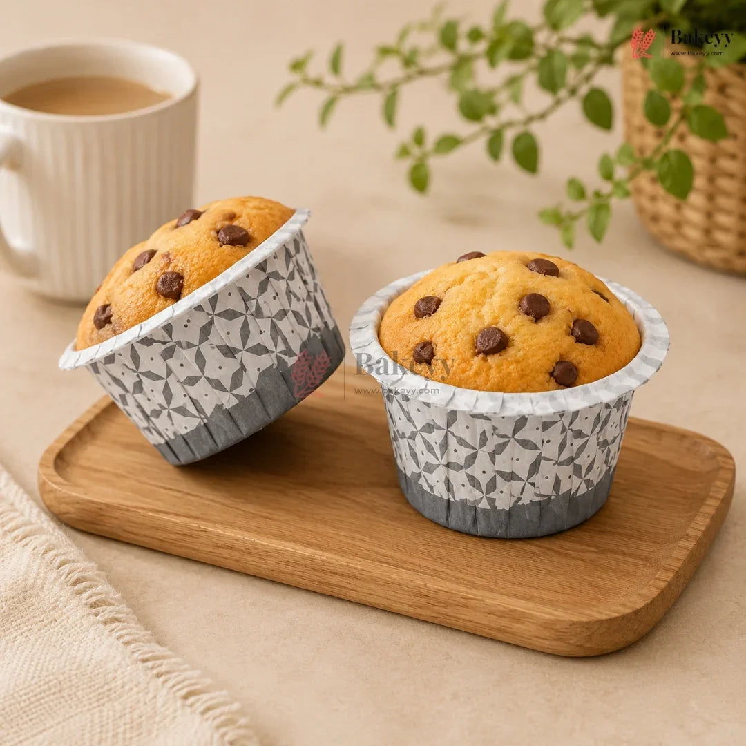 Two chocolate chip muffins in patterned paper cups on a wooden tray with a cup of coffee and plant in the background.