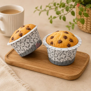 Two chocolate chip muffins in patterned paper cups on a wooden tray with a cup of coffee and plant in the background.