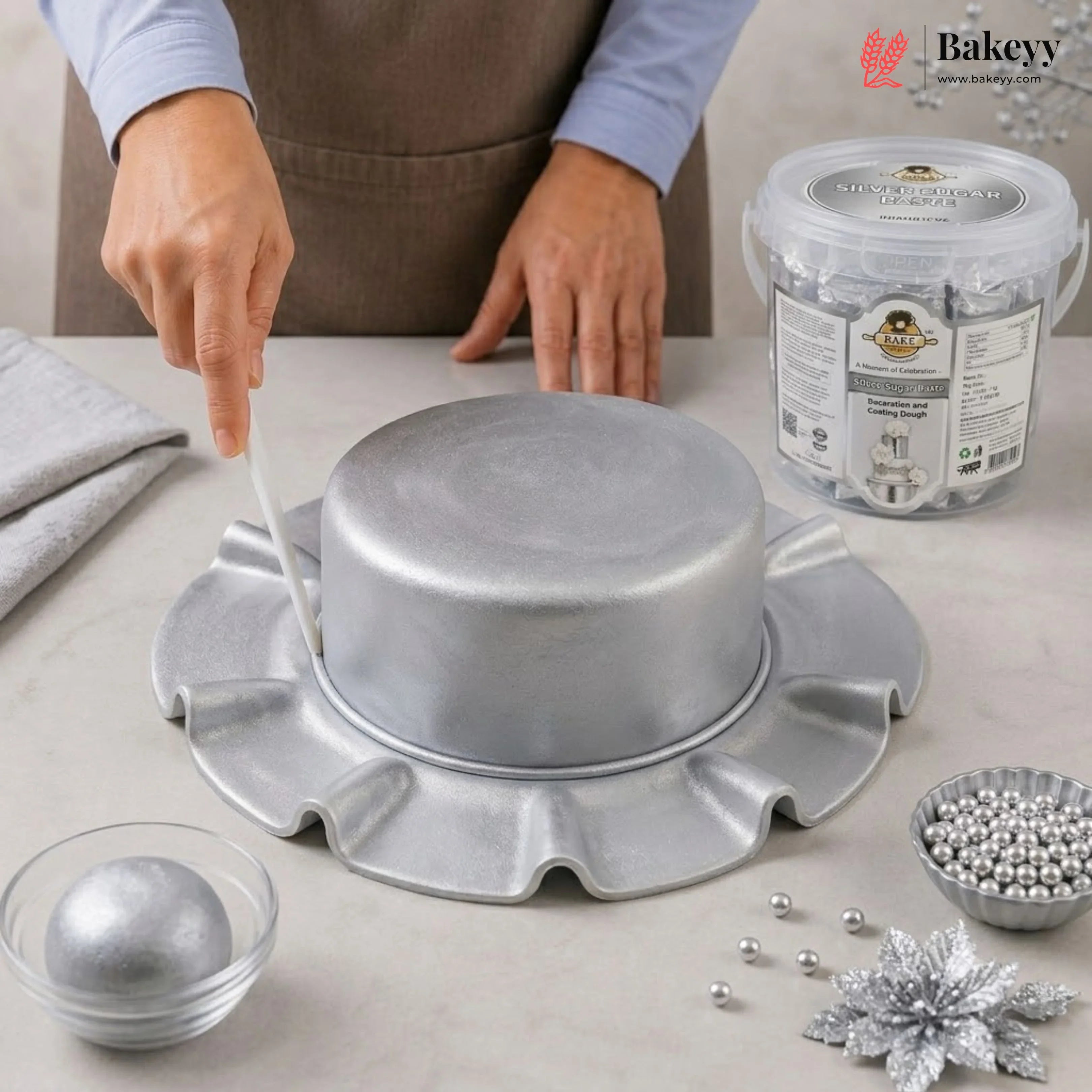 Person working with a silver cake stand and decorative items on a table, with Bakeyy branding visible.